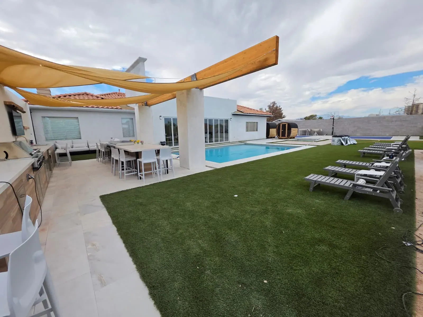 The backyard under its shade sails — pool, outdoor dining, loungers and the barrel saunas beyond