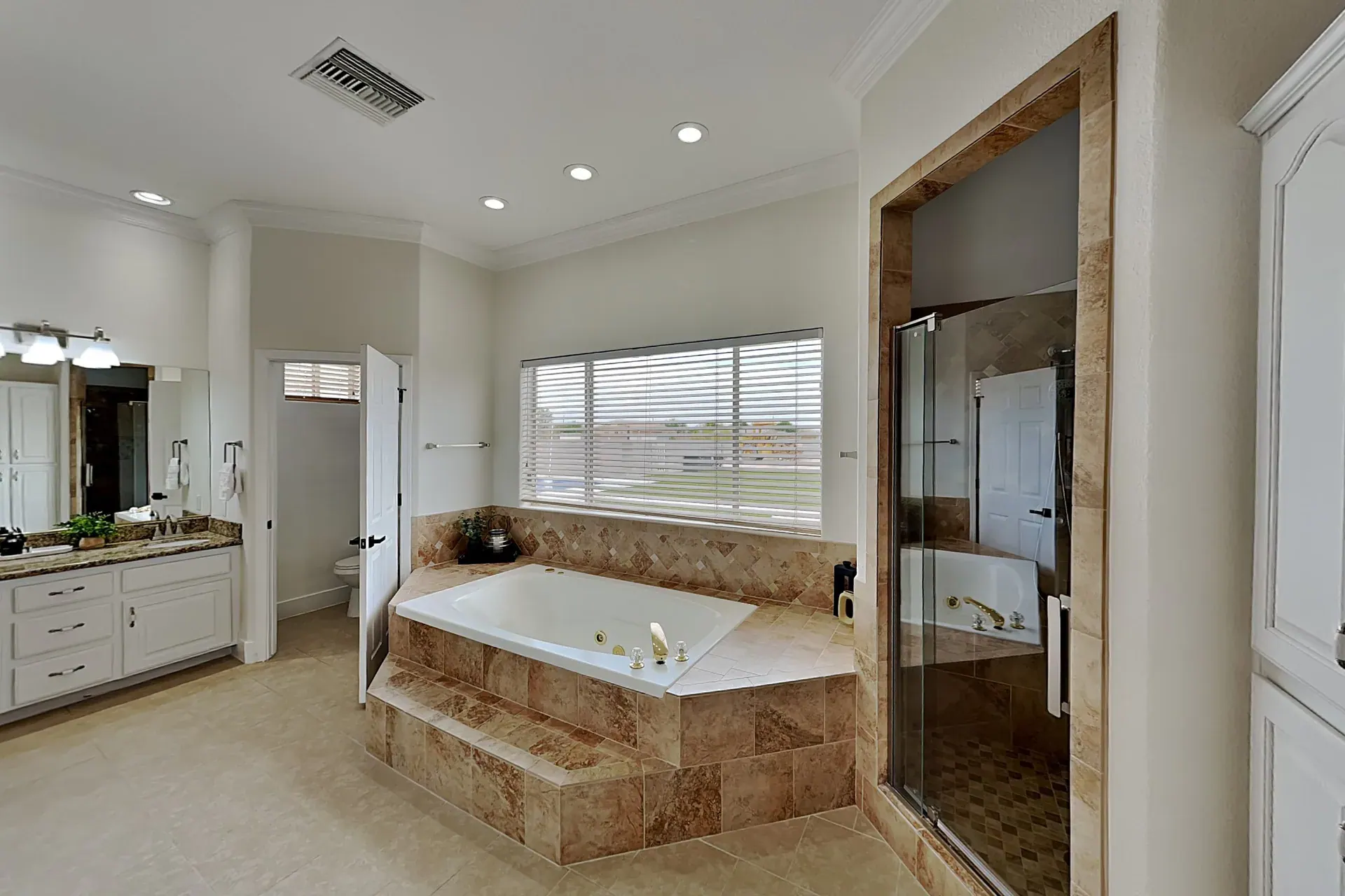 The largest bathroom — a jetted soaking tub set into travertine below a wide window, a double vanity, a separate glass shower and its own water closet