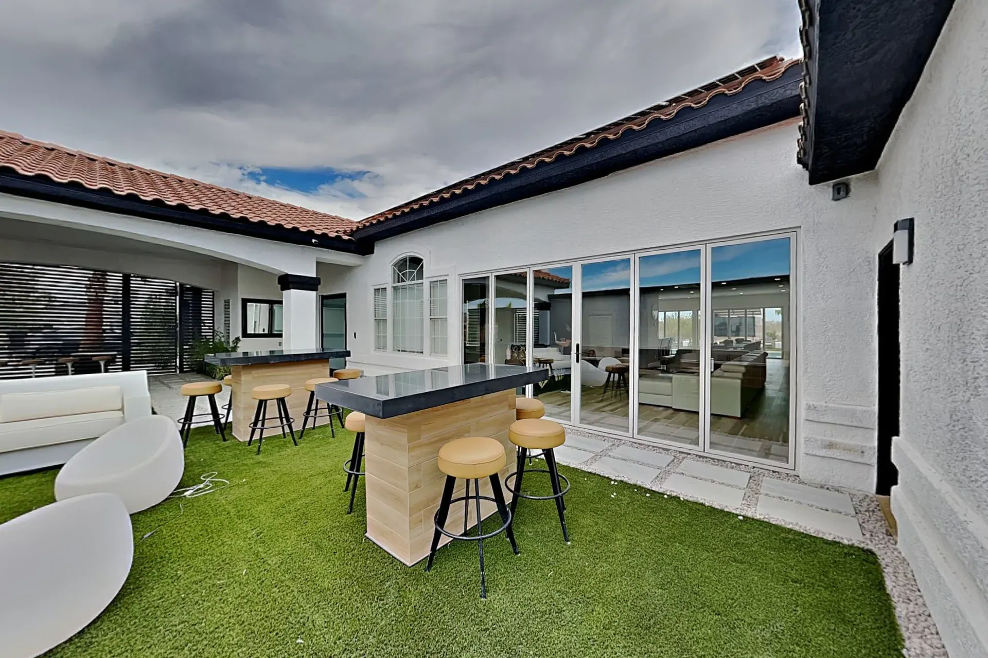 The courtyard on artificial turf — two high-top bar counters with cushioned stools and sculptural white chairs, opening onto the living room through a wall of glass