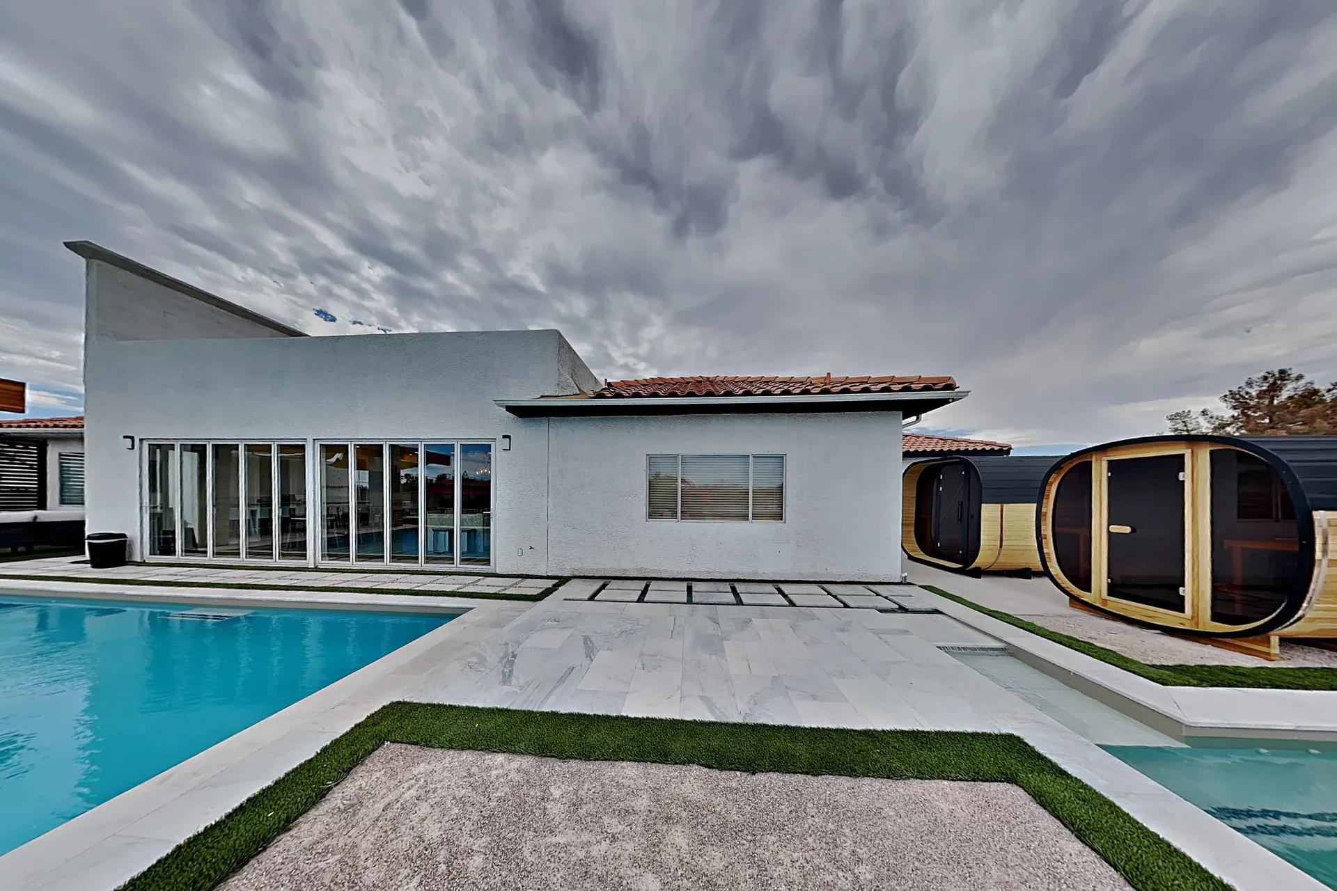 The rear of the villa under a dramatic sky, the pool in the foreground and the pair of cedar barrel saunas alongside