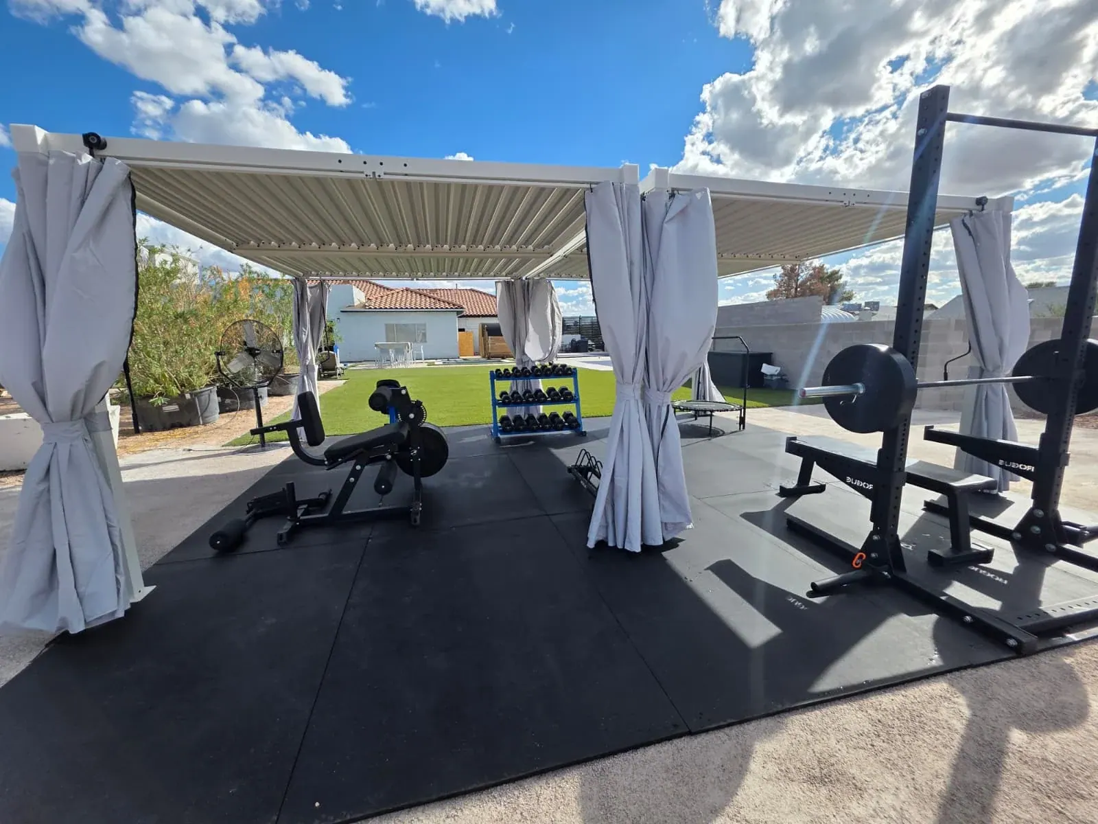 The outdoor gym under a louvered pergola — squat rack with barbell, benches, a leg machine and a dumbbell rack on rubber flooring