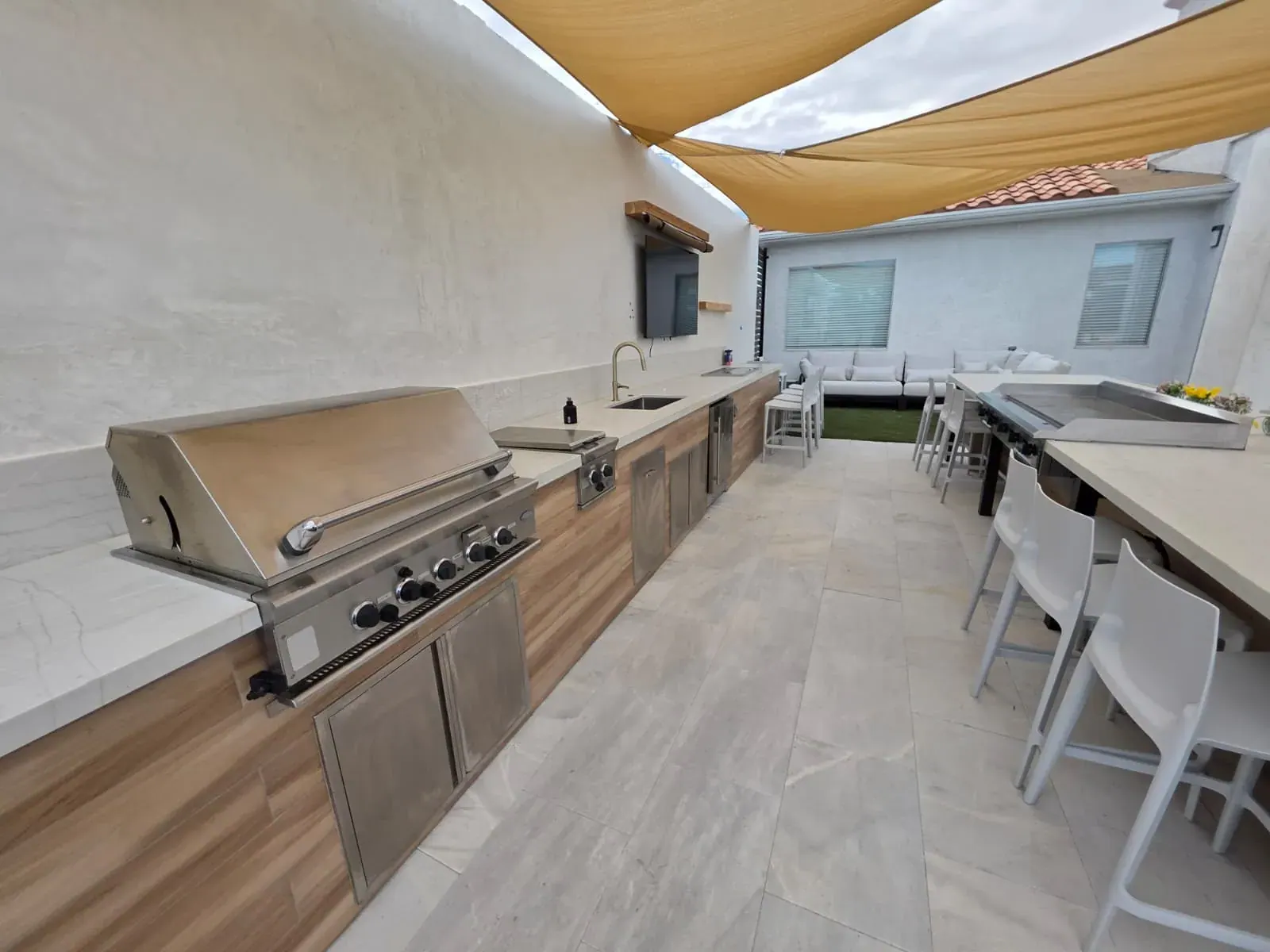 The outdoor kitchen under shade sails — built-in grill, side burners and a long stone counter with bar seating