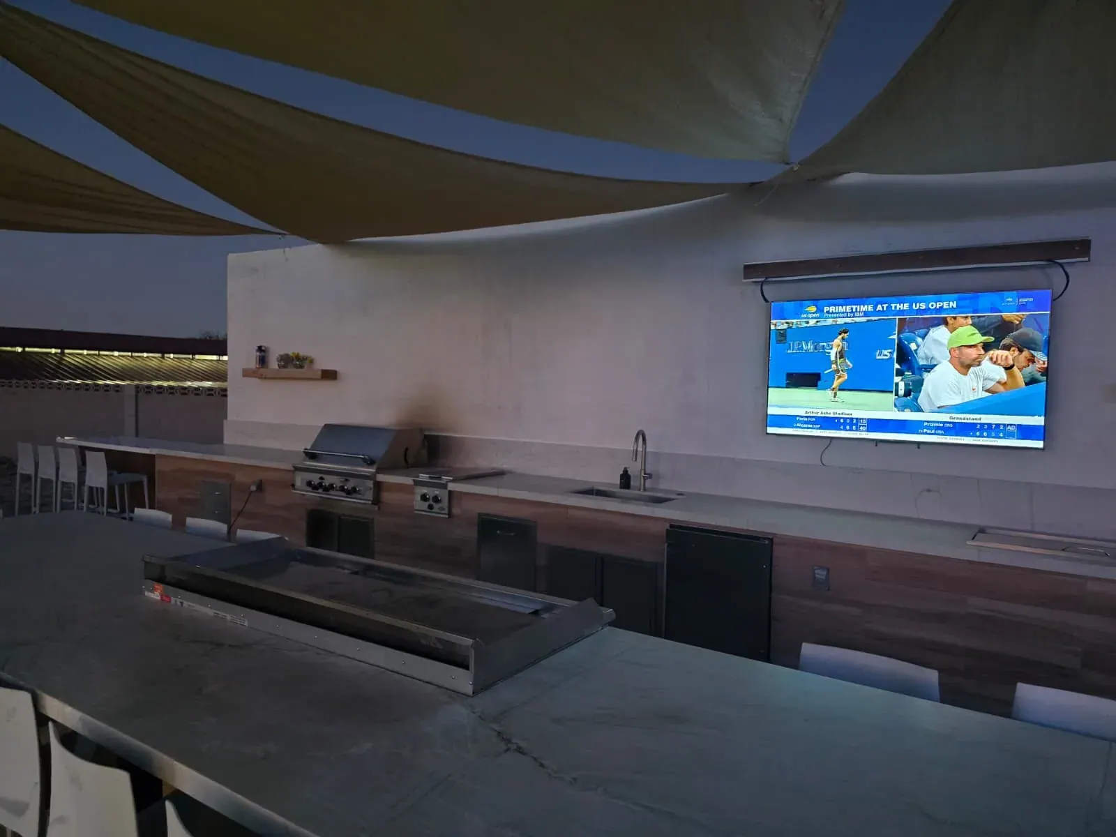 The outdoor kitchen after sunset — built-in grill, side burner, sink and refrigerator along a lit stone counter, with a TV mounted on the wall above