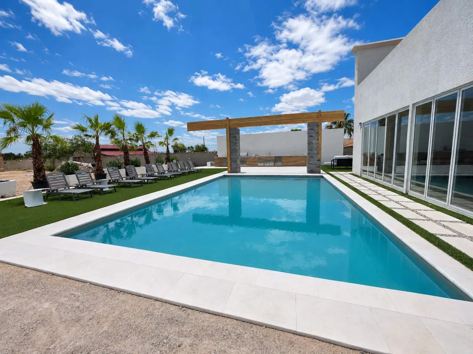 Heated pool lined with loungers and palm trees, with the pergola-covered outdoor kitchen beyond