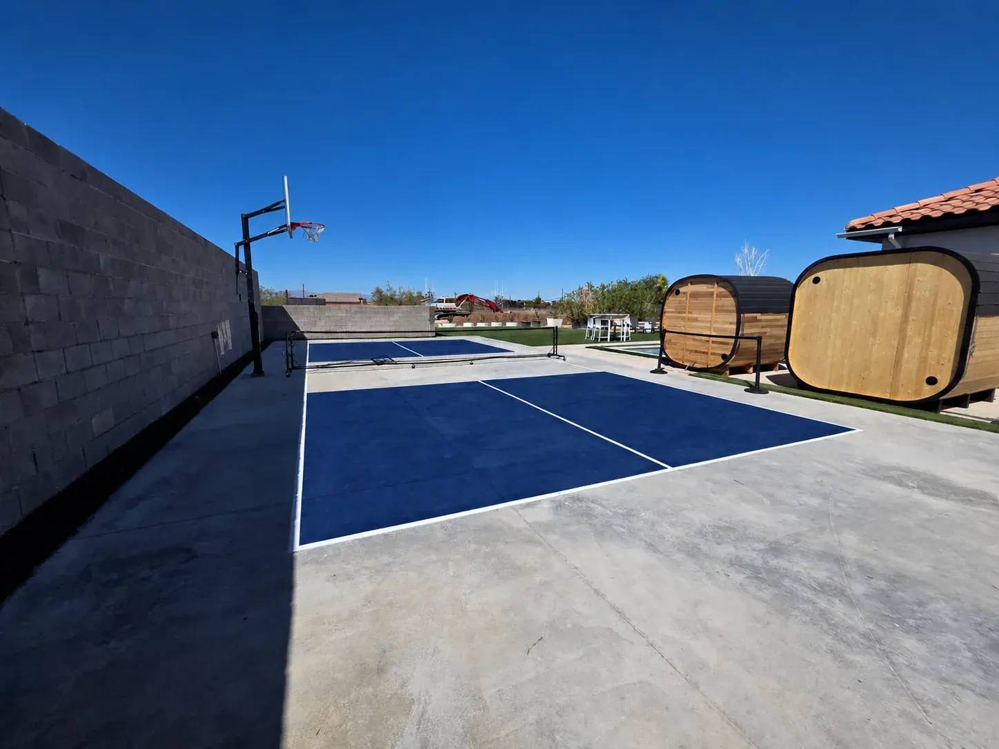 The private sports court — a blue pickleball court with net and a basketball hoop, the cedar barrel saunas along the far edge