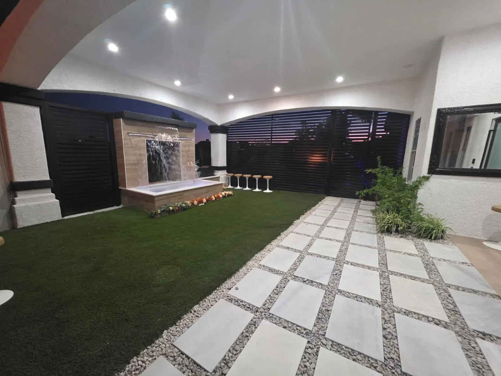 The front patio at dusk — a lit waterfall wall falling into the plunge basin, bar stools alongside and the sunset showing through the louvered privacy screens
